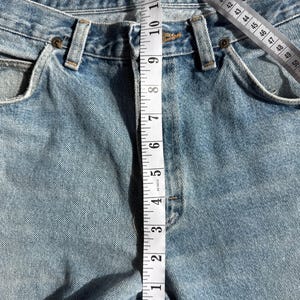 May include: Light blue denim jeans with a measuring tape showing a waist measurement. The tape reads approximately 11 inches. The jeans have classic five-pocket styling and a button and zipper closure.