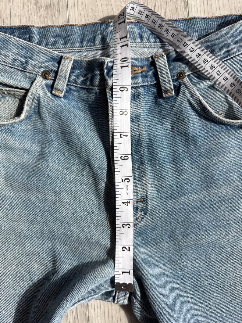 May include: Light blue denim jeans with a measuring tape showing a waist measurement. The tape reads approximately 11 inches. The jeans have classic five-pocket styling and a button and zipper closure.