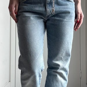 May include: Light blue denim jeans with a silver chain belt. The jeans have a classic five-pocket design and a slightly distressed look with a tear near the knee. The chain belt adds a touch of style.