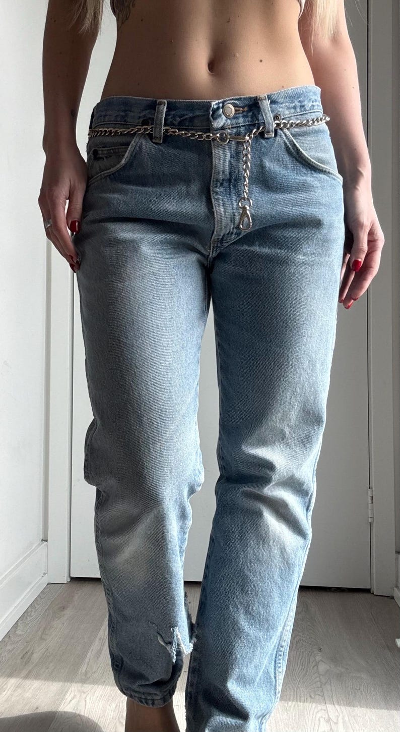 May include: Light blue denim jeans with a silver chain belt. The jeans have a classic five-pocket design and a slightly distressed look with a tear near the knee. The chain belt adds a touch of style.