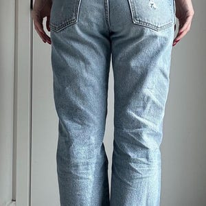 May include: Light blue denim jeans with a distressed look, featuring a ripped knee and a chain belt. The jeans have a classic five-pocket design and a faded wash, creating a vintage aesthetic.