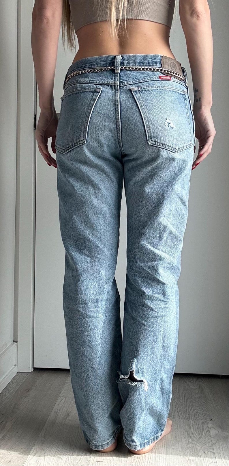 May include: Light blue denim jeans with a distressed look, featuring a ripped knee and a chain belt. The jeans have a classic five-pocket design and a faded wash, creating a vintage aesthetic.