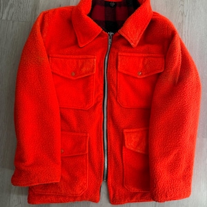 May include: Bright orange fleece jacket with a black and red plaid lining visible at the collar. The jacket has a full-length zipper, four front pockets, and a classic collar. A warm and stylish outerwear piece.