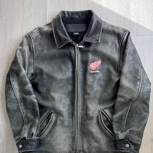Puede incluir: Chaqueta de cuero gris desgastado con cuello negro y cierre de cremallera. Presenta un logotipo de Toronto rojo y blanco en el pecho. Incluye dos bolsillos delanteros y puños abotonados. La chaqueta tiene un aspecto vintage y desgastado.