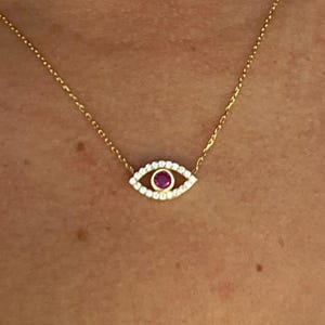 May include: Gold-toned necklace featuring an eye-shaped pendant. The pendant has a ruby center, surrounded by a halo of small, clear gemstones. The necklace is delicate and appears to be worn around the neck.