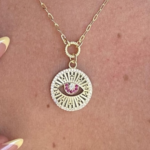 May include: Gold-toned necklace featuring a circular pendant with an eye design. The pendant has a pink center and is surrounded by clear stones. The chain is delicate and the necklace is worn on a person's neck.