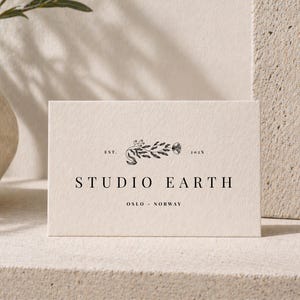 May include: A rectangular card with the words "STUDIO EARTH" in a serif font. The card has a floral design above the text and the words "OSLO - NORWAY" below. The card is cream-colored and sits on a light-colored surface.