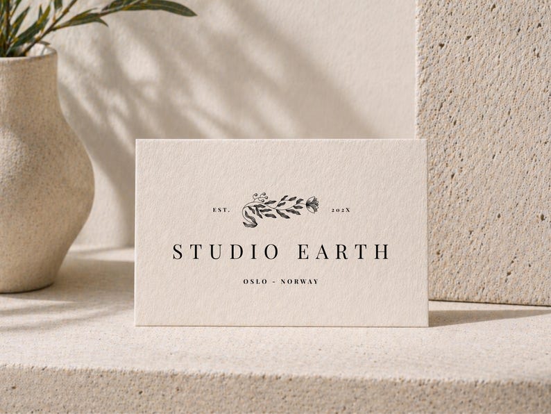 May include: A rectangular card with the words "STUDIO EARTH" in a serif font. The card has a floral design above the text and the words "OSLO - NORWAY" below. The card is cream-colored and sits on a light-colored surface.