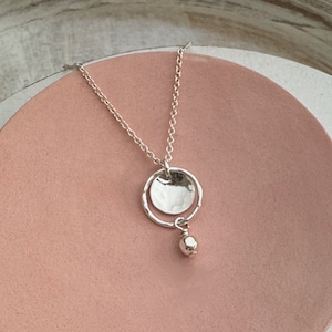 May include: A silver necklace with a hammered disc pendant and a small dangling bead. The necklace is displayed on a pink, circular plate, set against a neutral background. The pendant has a textured surface.