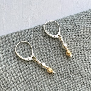 May include: A pair of dangle earrings with silver lever-back closures. Each earring features a series of small silver beads and gold-colored beads. The earrings are displayed on a gray, textured surface.