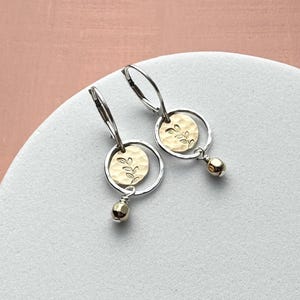 May include: A pair of dangle earrings featuring a silver hoop attached to a lever back. Inside each silver hoop hangs a small, round, hammered gold-tone disc engraved with a delicate leaf sprig design, accented by a tiny, polished gold-tone bead.