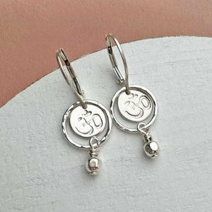 May include: A pair of silver earrings featuring a circular design with the Om symbol. Each earring has a small, dangling bead and a lever-back closure. The earrings are displayed on a white surface with a soft pink background.