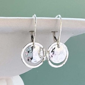 May include: A pair of silver dangle earrings. Each earring features a hammered, circular disc within a silver hoop. The earrings are displayed on a white surface against a muted teal background. The earrings are designed to catch the light.