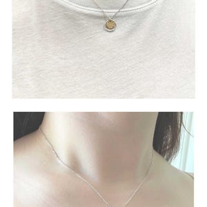 May include: A silver necklace with a small, round pendant. The pendant has a textured surface and a silver border. The necklace is worn against a white t-shirt. The image also shows the necklace on a person's neck.