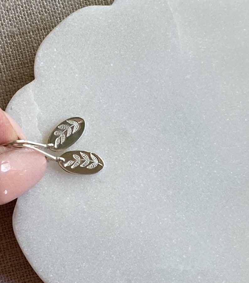 Op de afbeelding: Een paar zilverkleurige ovale oorbellen. Elke oorbel heeft een delicaat bladontwerp. De oorbellen worden getoond tegen een witte marmeren ondergrond, die hun ingewikkelde details en vakmanschap benadrukt. Een klassiek sieraad.