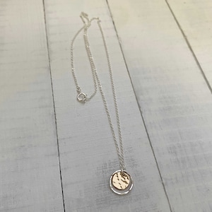 May include: A silver necklace with a delicate chain and a circular pendant. The pendant features a gold-colored hammered disc inside a silver ring. The necklace is displayed on a white wooden surface.