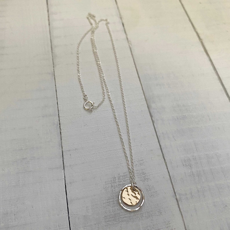 May include: A silver necklace with a delicate chain and a circular pendant. The pendant features a gold-colored hammered disc inside a silver ring. The necklace is displayed on a white wooden surface.
