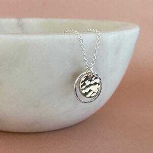 May include: A delicate silver necklace featuring a hammered gold-tone disc pendant suspended within a silver ring. The necklace has a fine chain and is displayed against a white background with a soft pink backdrop. The pendant has a textured surface.