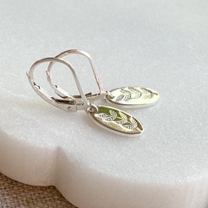 Op de afbeelding: Een paar zilveren oorhangers met oorhaken. Elke oorbel heeft een ovale bedel met een gedetailleerd bladerontwerp. De oorbellen worden gepresenteerd op een wit marmeren oppervlak.