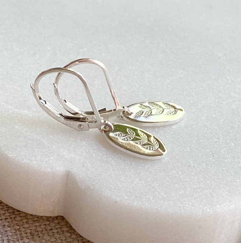 Op de afbeelding: Een paar zilveren oorhangers met oorhaken. Elke oorbel heeft een ovale bedel met een gedetailleerd bladerontwerp. De oorbellen worden gepresenteerd op een wit marmeren oppervlak.