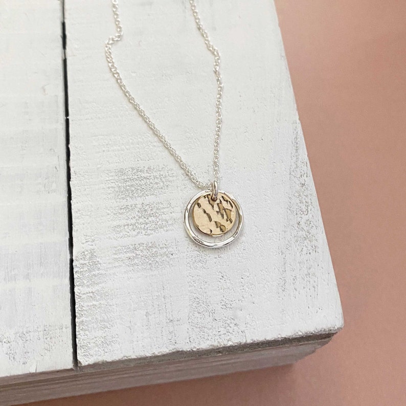 May include: A silver necklace with a delicate chain. The pendant features a gold-colored, textured disc inside a silver ring. The necklace is displayed on a white wooden surface against a soft pink background.