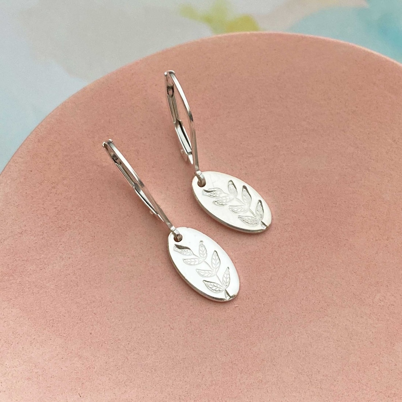 May include: Silver hoop earrings with oval shaped charms. Each charm has a detailed leaf design.