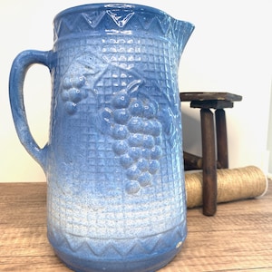 May include: A blue and white glazed ceramic pitcher with a grape design. The pitcher has a handle and a wide mouth.