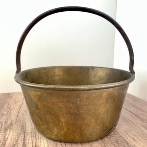 May include: A vintage brass cauldron with a dark metal handle. The cauldron has a wide, rounded body and a slightly flared rim. The brass has a warm, aged patina, and the handle arches over the top.