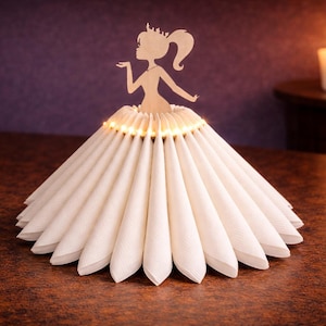 Pode incluir: Um porta-guardanapos decorativo em forma de princesa com um vestido. O vestido é feito de guardanapos brancos dobrados, e a silhueta da princesa é feita de madeira castanha clara. Pequenas luzes estão em volta da cintura da princesa.
