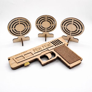 May include: Wooden toy gun with the text "PEW PEW!" and three wooden target practice boards. The gun has a textured grip and a yellow elastic band. The targets have concentric circles.