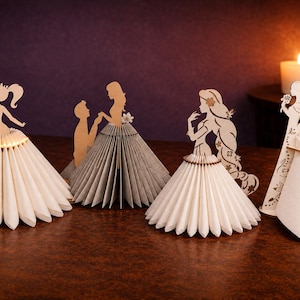 Pode incluir: Quatro centros de mesa decorativos com silhuetas de madeira cortadas a laser presas a saias de papel plissado. As figuras incluem uma princesa, um casal, uma personagem de cabelo comprido e uma figura em pé, com uma saia iluminada por pequenas luzes.