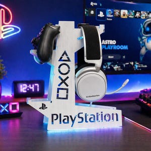 May include: A white PlayStation gaming headset and controller stand. The stand features the PlayStation logo and controller symbols. A black controller and white headset are displayed on the stand. The background includes a monitor and neon PlayStation logo.