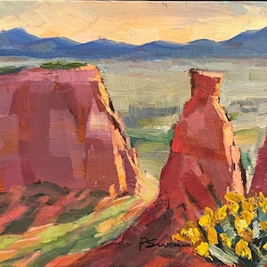 Puede incluir: Pintura al óleo de un paisaje desértico con formaciones rocosas rojas y flores silvestres amarillas. El cielo es una mezcla de amarillo pálido y azul, con montañas azules al fondo. La firma del artista está en la esquina inferior derecha.