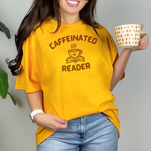 Op de afbeelding: Een geel T-shirt met de tekst "caffeinated reader" in bruine letters. Het ontwerp bevat een gestileerde afbeelding van een kopje koffie met stoom die eruit opstijgt en een boek eronder.
