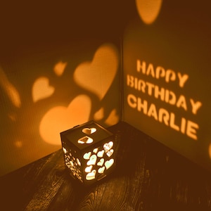 May include: A wooden cube-shaped lamp with heart cutouts, casting heart-shaped shadows on the wall. The words "HAPPY BIRTHDAY CHARLIE" are projected on the wall. The lamp emits a warm, orange glow.