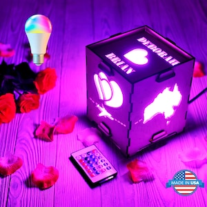 May include: A wooden cube night light with a remote control. The cube has two hearts, a plane, and a map of Australia on it. The names "Brian" and "Deborah" are engraved on the cube. The light is on and glowing purple.