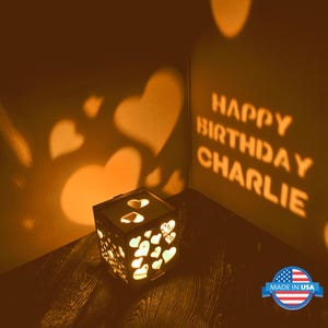 May include: A wooden shadow box with a light inside that projects the words "Happy Birthday Charlie" and hearts onto the wall. The box is made of dark wood and has a light brown finish. The box is sitting on a wooden table. The box is made in the USA.