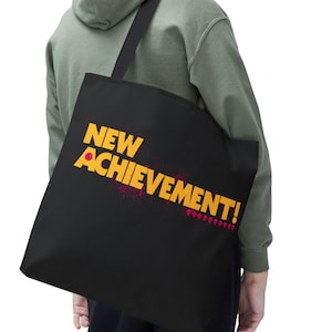 May include: Black tote bag featuring the text "NEW ACHIEVEMENT!" in yellow and red. The bag is slung over the shoulder. The person is wearing a green hoodie.