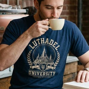 Pode incluir: Uma pessoa num café, vestindo uma camiseta azul marinho com a inscrição "Luthadel University", bebe de uma caneca bege. A pessoa está sentada em uma mesa de madeira com um livro. O fundo apresenta tijolos expostos e luminárias suspensas.