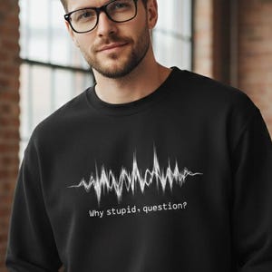 May include: Black sweatshirt with a white graphic of a sound wave and the text "Why stupid, question?" The sweatshirt is a crew neck style and is made of a soft material. The person in the image is wearing black glasses.