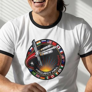 Peut inclure: T-shirt blanc à col et manches noirs. Le t-shirt présente un motif circulaire avec un dessin de navette spatiale, le texte "PROJECT HAIL MARY" et des drapeaux de différents pays.