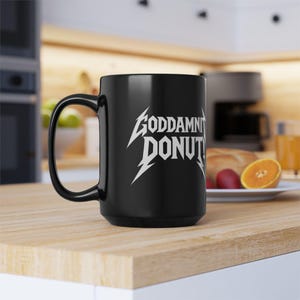 Black ceramic mug with bold white text in a Metallica-style font that reads “Goddamnit Donut,” inspired by Dungeon Crawler Carl. The mug is set on a modern kitchen counter, perfect for fans of LitRPG, Princess Donut, and Matt Dinniman's cult book series. Ideal for coffee or tea lovers who enjoy a mix of humor, chaos, and fandom in their daily routine.