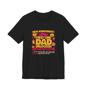 Puede incluir: Camiseta negra con un gráfico colorido que dice "NEW ACHIEVEMENT! FIRST TIME DAD UNLOCKED! HAPPY FIRST FATHER'S DAY!". El diseño incluye un trofeo, un bigote, un biberón y otros iconos relacionados con bebés. La camiseta también enumera los efectos secundarios y las recompensas.