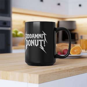 Black ceramic mug with bold white text in a Metallica-style font that reads “Goddamnit Donut,” inspired by Dungeon Crawler Carl. The mug is set on a modern kitchen counter, perfect for fans of LitRPG, Princess Donut, and Matt Dinniman's cult book series. Ideal for coffee or tea lovers who enjoy a mix of humor, chaos, and fandom in their daily routine.
