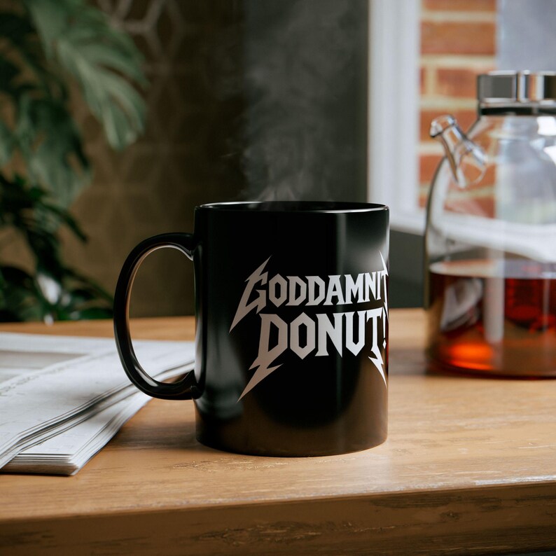 Black ceramic mug with bold white text in a Metallica-style font that reads “Goddamnit Donut,” inspired by Dungeon Crawler Carl. The mug is set on a modern kitchen counter, perfect for fans of LitRPG, Princess Donut, and Matt Dinniman's cult book series. Ideal for coffee or tea lovers who enjoy a mix of humor, chaos, and fandom in their daily routine.