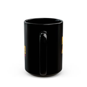 May include: A black ceramic coffee mug with a large handle. The mug has yellow and pink text on the side. The mug is a standard size for hot beverages.
