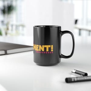 May include: A black ceramic mug with a large handle. The mug features the word "MENT!" in yellow block letters, with a row of pink skulls below. The mug is on a white surface.