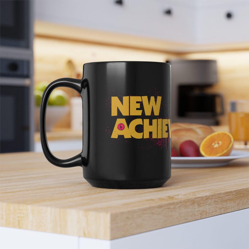 May include: A black ceramic mug with a large handle. The mug features the words "NEW ACHIE" in yellow block letters with a pink accent. The mug is sitting on a light-colored wooden surface.