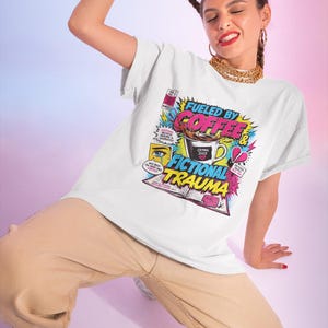 Woman posing dynamically wearing white Fueled by Coffee and Fictional Trauma bookish graphic tee, colorful comic print, pink blue gradient background, funny reader shirt