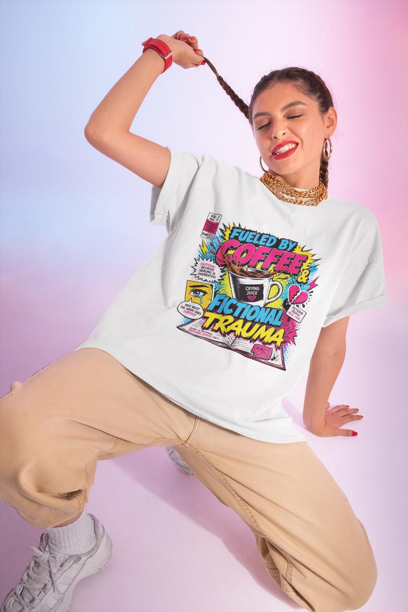 Woman posing dynamically wearing white Fueled by Coffee and Fictional Trauma bookish graphic tee, colorful comic print, pink blue gradient background, funny reader shirt